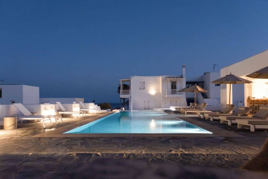 A serene evening poolside scene at a boutique hotel with modern white architecture and soft outdoor lighting. The glowing pool reflects the peaceful ambiance, surrounded by sun loungers and umbrellas.