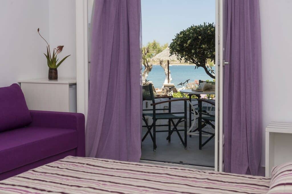 A cozy bedroom with a purple sofa and curtains, opening onto a patio with a dining setup, shaded by trees, overlooking the clear blue sea in the distance.