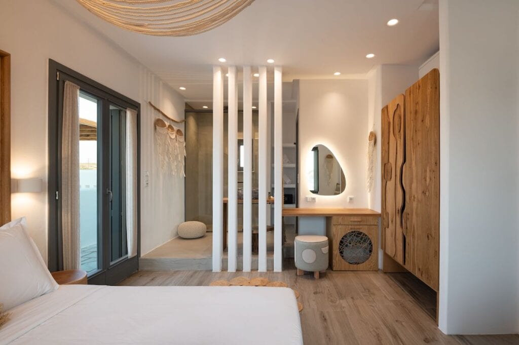 A stylish suite blending natural wood elements with modern aesthetics, featuring a cozy bed, an open-concept bathroom with decorative lighting, and a private terrace with sliding glass doors.