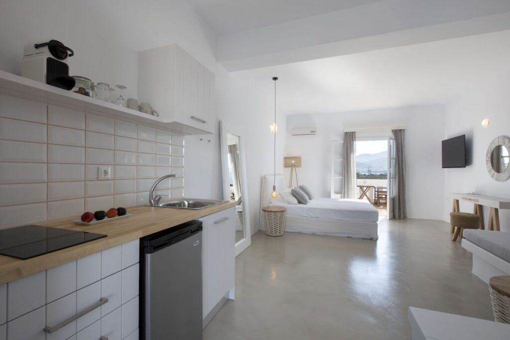 A modern, airy studio apartment at Kouros Village with an open-plan layout, featuring a kitchenette, a bed, and a private balcony overlooking the sea. The neutral decor, natural light, and minimalist furnishings create a tranquil and inviting space.