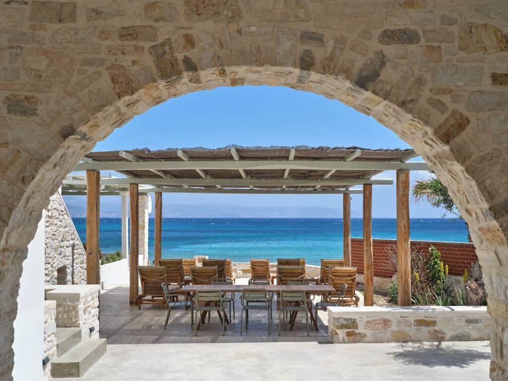 A Mediterranean-style outdoor dining area with rustic stone archways, wooden tables and chairs, shaded by a pergola, all facing the shimmering turquoise sea under a bright blue sky.