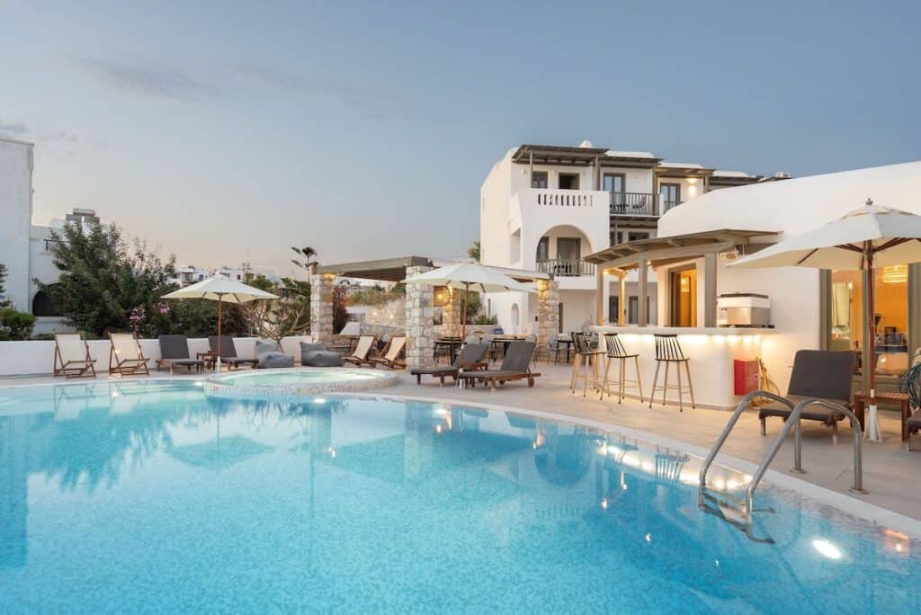 A dreamy poolside view at dusk, featuring a luxurious hotel with white Cycladic architecture. The large swimming pool is surrounded by sun loungers, umbrellas, and a chic outdoor bar. Warm lighting highlights the elegant, relaxing atmosphere.