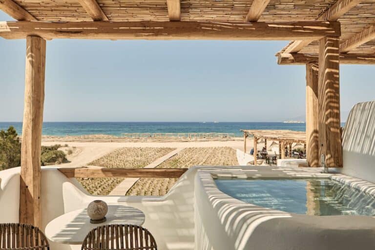 A bohemian-style hotel balcony with a built-in jacuzzi overlooking the ocean. A small table with rattan chairs is set under a wooden pergola, providing a perfect spot to soak in the sea breeze. The natural tones of the decor blend seamlessly with the surrounding landscape.