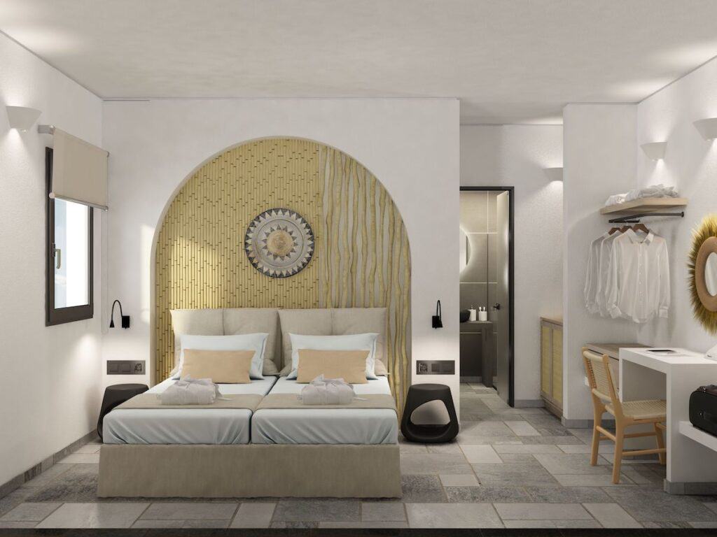 A stylish bedroom featuring a neutral color palette with a bamboo-textured arched headboard, a cozy double bed with soft linens, and a minimalistic workspace. The room includes an open wardrobe and leads to a sleek modern bathroom.