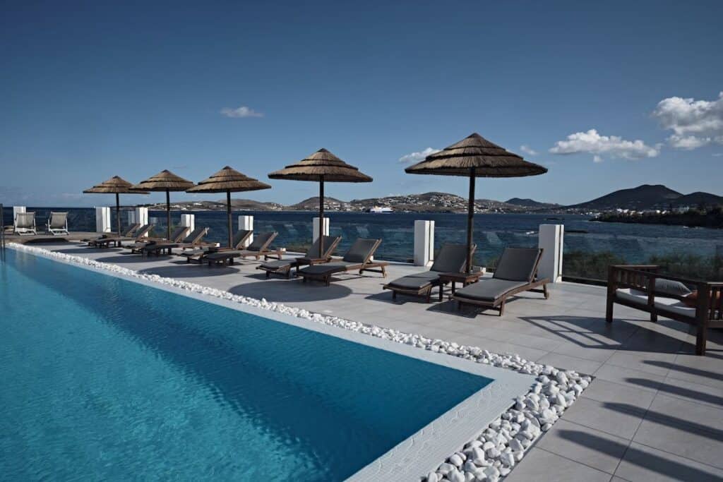 A serene infinity pool lined with sun loungers and thatched umbrellas, overlooking the deep blue sea. The scene captures a peaceful ambiance with distant islands and hills under a clear sky.