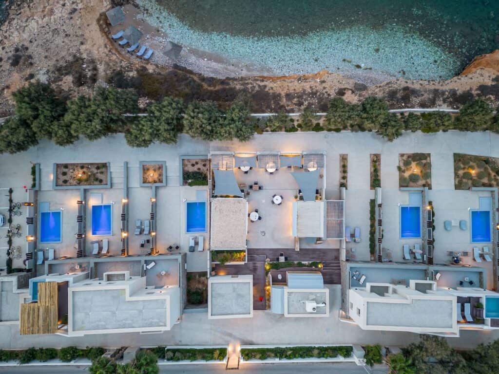 An aerial view of a beachfront resort with modern architecture, featuring multiple private pools, sun loungers, and lush greenery. The property is directly adjacent to a rocky shoreline with crystal-clear waters.