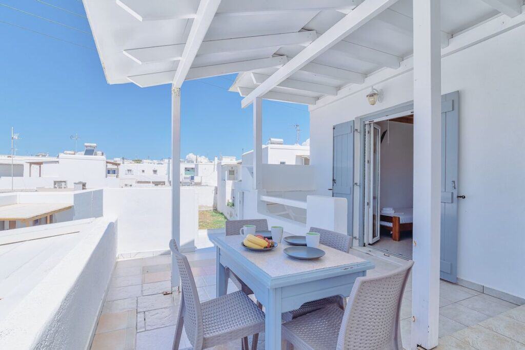A sunlit balcony with a table and chairs at Pavlos Place in Paros, set up for an al fresco breakfast with fruit and coffee cups. White-washed buildings and a bright blue sky in the background create a classic Greek island atmosphere.