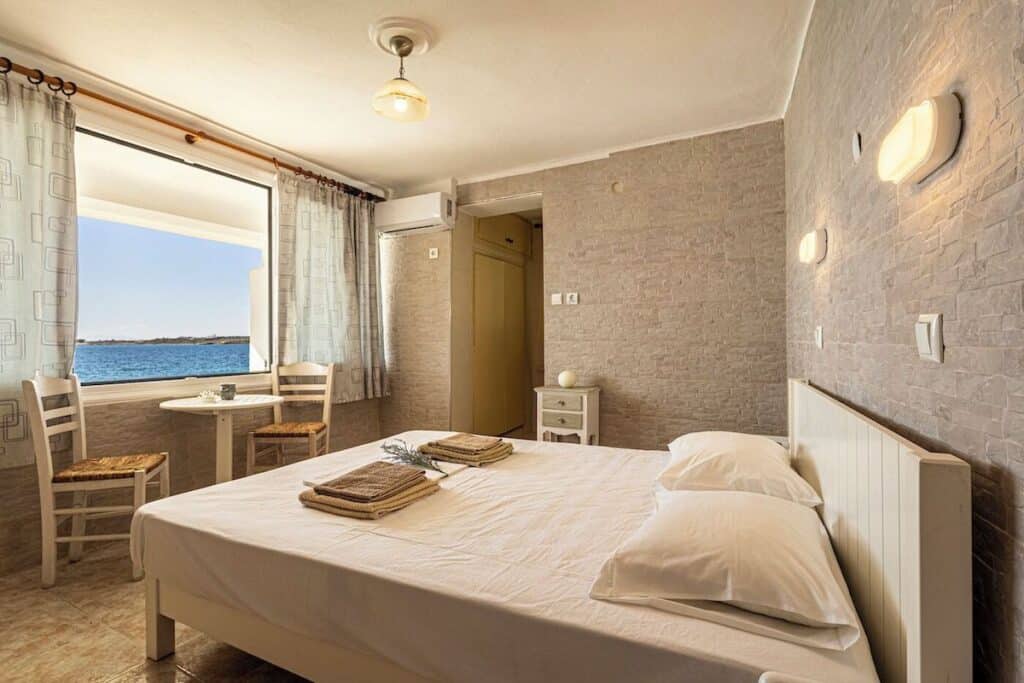 A cozy seaside bedroom featuring a simple white bed with fresh linens, a rustic stone-textured wall, and a small dining area by a large window that offers stunning views of the blue sea.