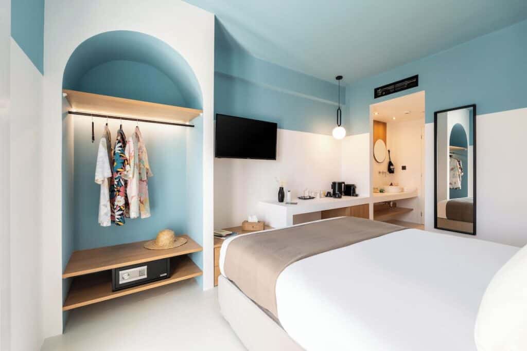A room at Play Hotel in Paros with a sleek blue-and-white color scheme, featuring a neatly made bed, a stylish open wardrobe with colorful clothes hanging, and a mounted flat-screen TV. A bathroom with a large mirror is visible in the background.