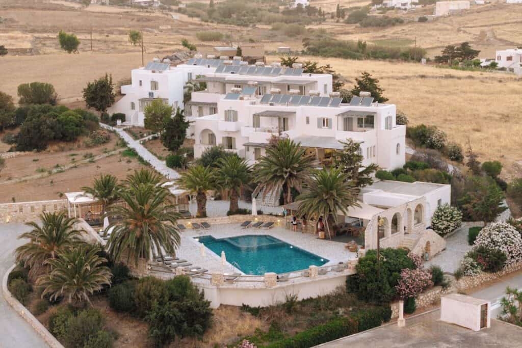 Pnoi Hotel complex with white Mediterranean-style buildings, surrounded by lush greenery and palm trees. The central courtyard features a large swimming pool with sun loungers, creating a relaxing vacation setting