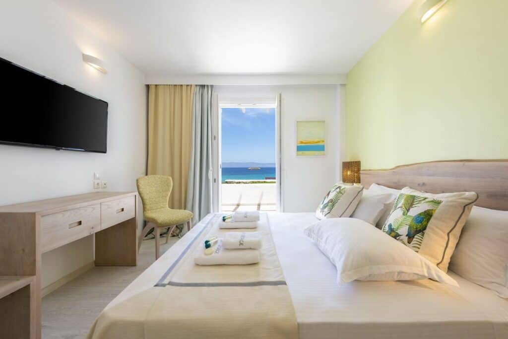 A bright and airy beachfront hotel room with a neatly made bed, a flat-screen TV, and a wooden desk. The open balcony door reveals a stunning sea view with blue waters and a clear sky.