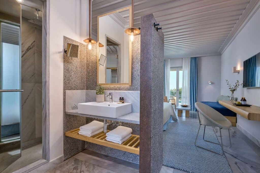 A contemporary suite with a semi-open bathroom featuring marble and pebble textures, a spacious vanity, and a stylish bedroom with ocean-inspired tones and modern decor.