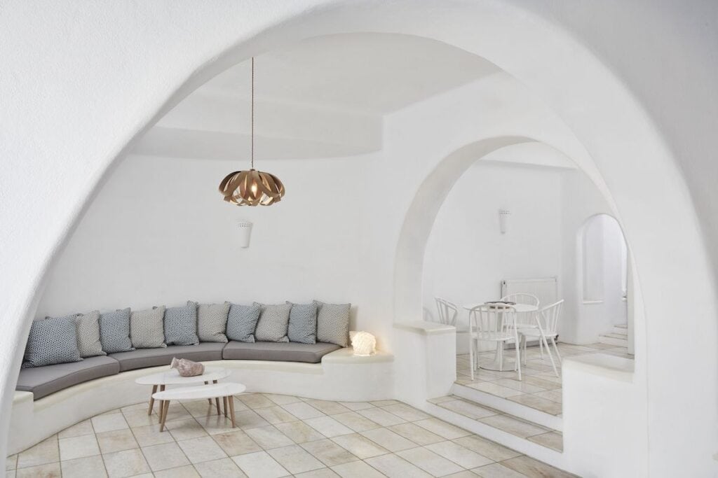 A bright, open lounge area with arched doorways, built-in white seating adorned with neutral-colored cushions, and a stylish pendant light and minimalist decor.