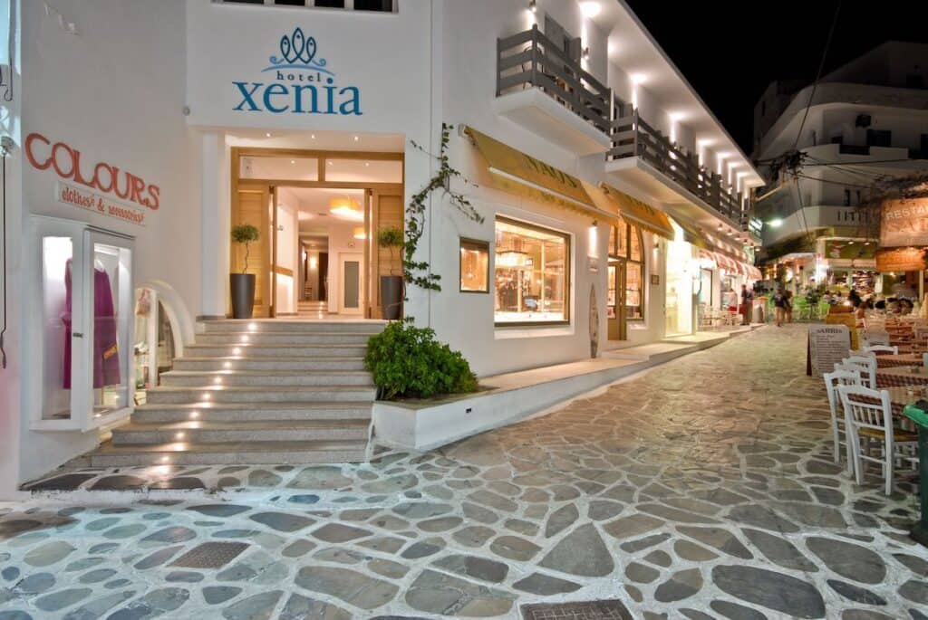 A lively nighttime scene outside Xenia Hotel, with an illuminated entrance leading to a stylish lobby. The hotel is situated along a cobblestone pedestrian street lined with shops, cafes, and vibrant nightlife.