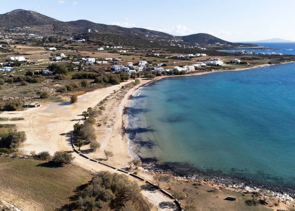 A bird's eye view of Piso Aliki Beach, with it's calm water, small beach, and sandy parking area separated by trees. There are lots of houses and farmland in the landscape and multiple other coves in the distance.