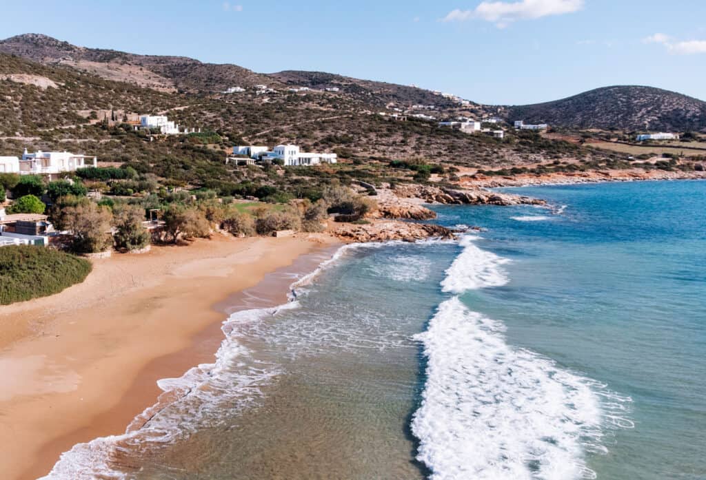 Faragas Beach on a relaxing day in Paros, with waves slowly crashing into the sandy shore lined with vegetation. You can also see some whitewashed buildings and rolling hills in the distance. 