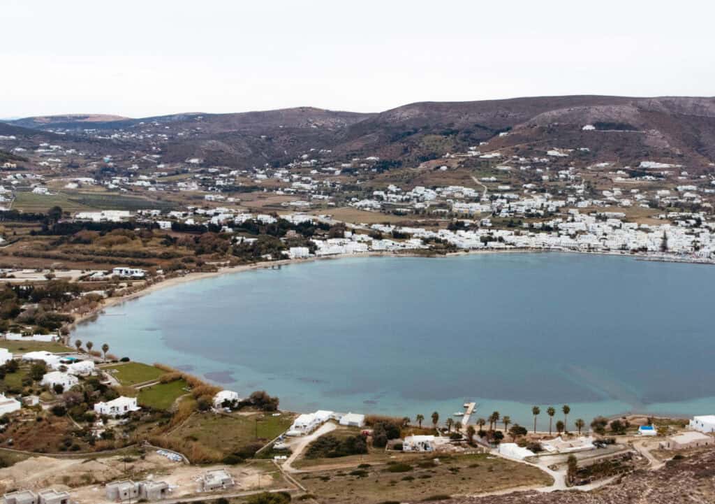 Livadia Beach in the bay next to Parikia Town. There's a small dock without boats and sparse palm trees lining the bay, with a sandy beach starting just at the edge of the town. 
