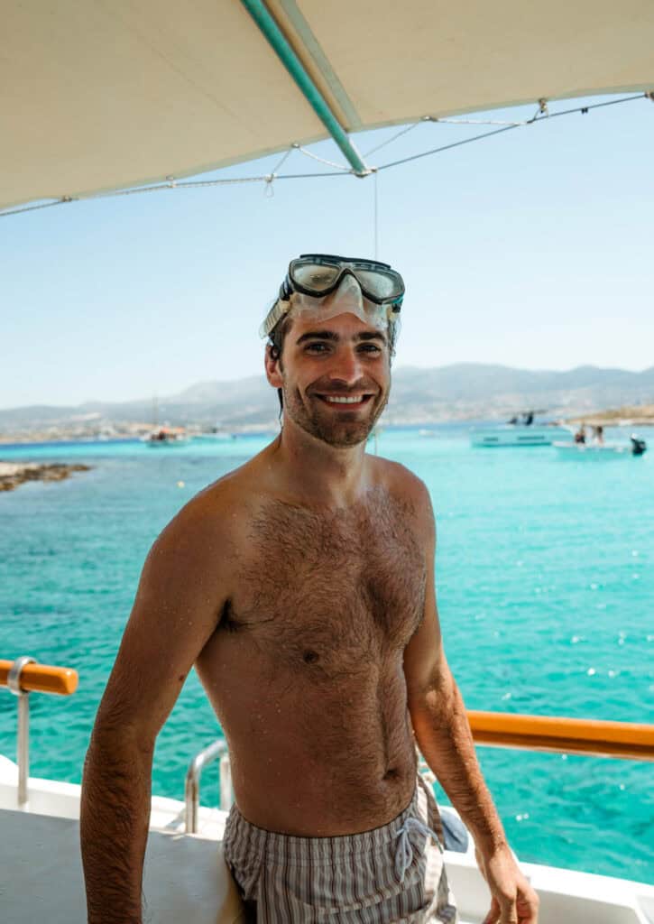 Caleb wearing swim goggles on a boat during a sunny Paros boat trip, with crystal-clear waters and distant hills in the background.