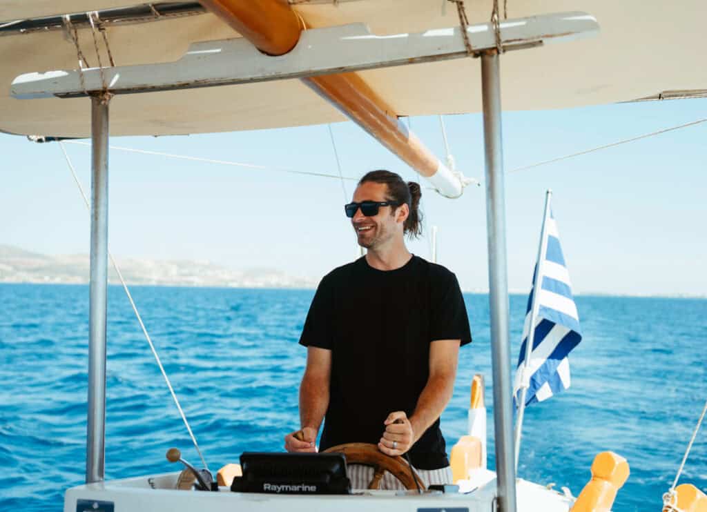 Caleb at the helm of the boat, smiling under the sun with a Greek flag waving behind.