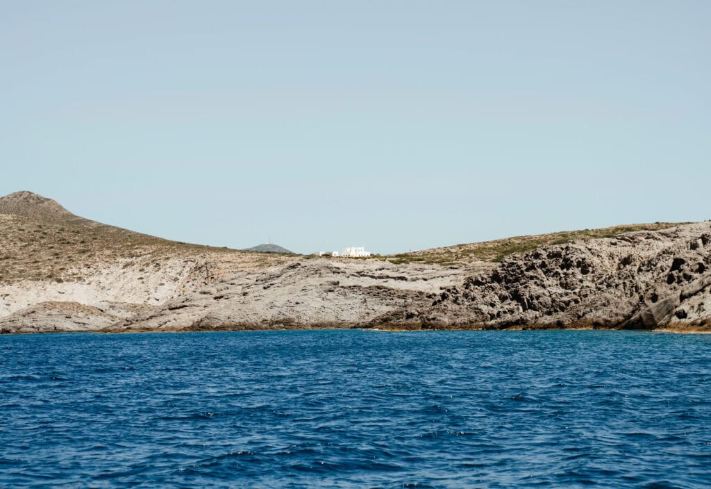 Coastal view of rocky terrain and dry hills under a pale blue sky, with a white building visible in the distance above the deep blue sea in Paros