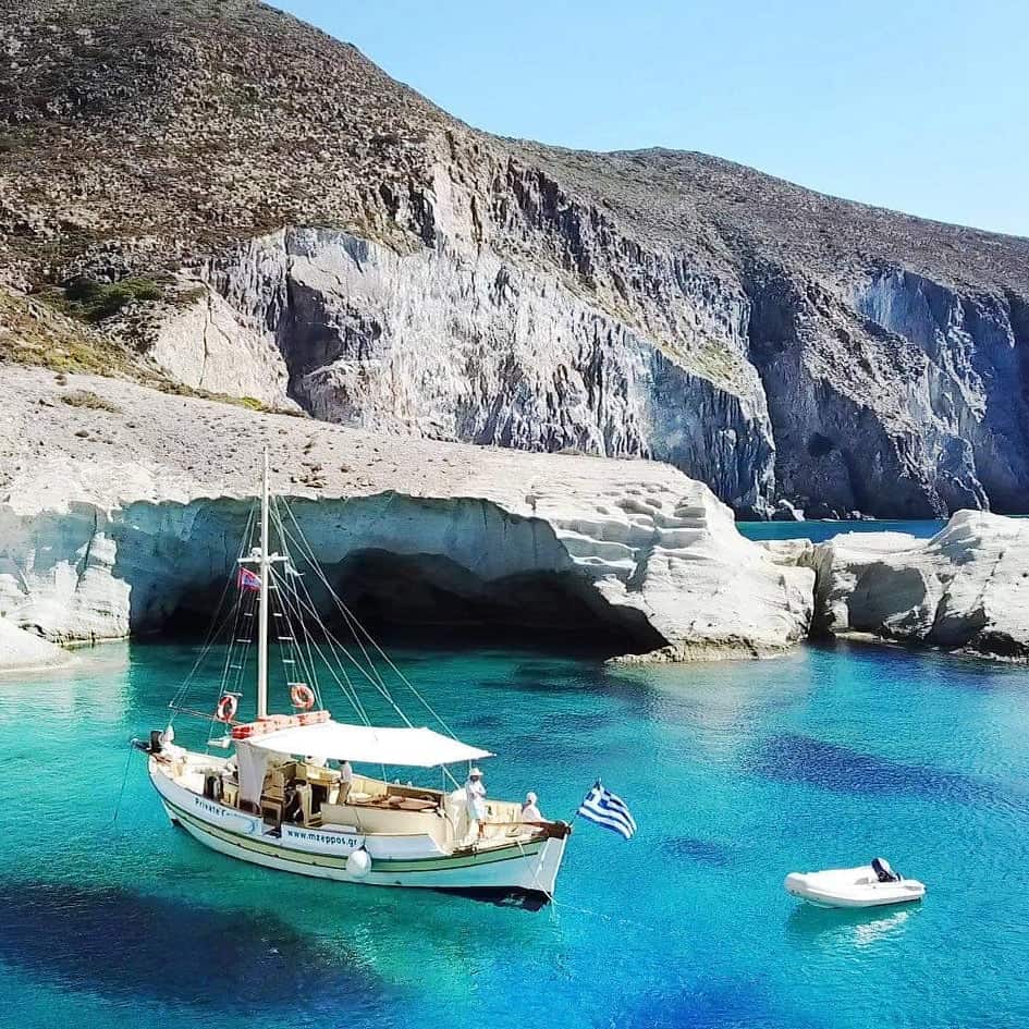 Traditional wooden boat anchored in crystal-clear turquoise waters surrounded by white sculpted cliffs in Paros, Greece. This boat tour in Paros offers visitors a chance to explore hidden coves and experience the island’s stunning coastline.