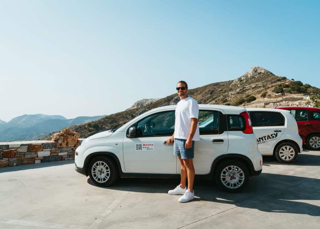 Caleb standing beside a white rental car in a mountain parking area on Naxos island, highlighting the freedom and accessibility that comes with renting a car in Naxos