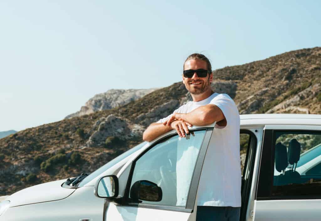 Caleb smiling beside a white rental car with scenic Naxos mountains in the background, after renting a car in Naxos to explore the island