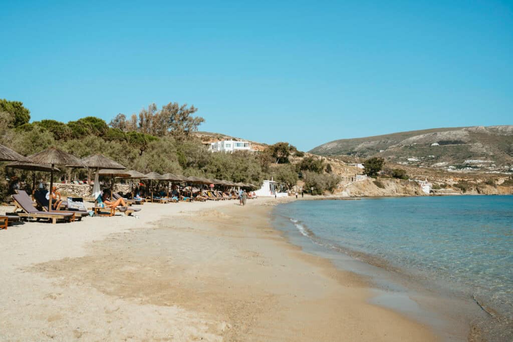 View of the quieter end of Marcello Beach Paros with rustic wooden umbrellas, gentle waves, and distant views of the Parikia hillside.