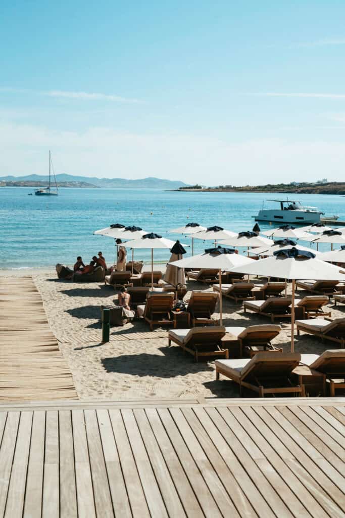 Boardwalk leading to umbrella-covered loungers at Marcello Beach Paros, with boats anchored in the bay and views of nearby islands