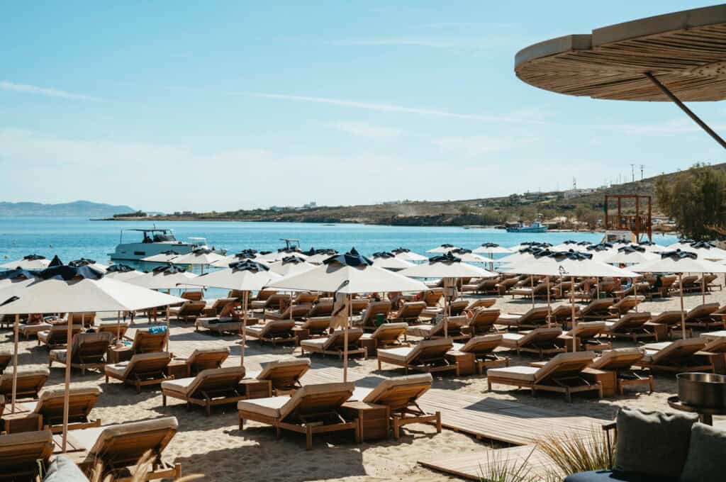 Rows of beach loungers and umbrellas facing the sea at Marcello Beach Paros, with yachts anchored offshore and a wooden lifeguard tower in the distance.