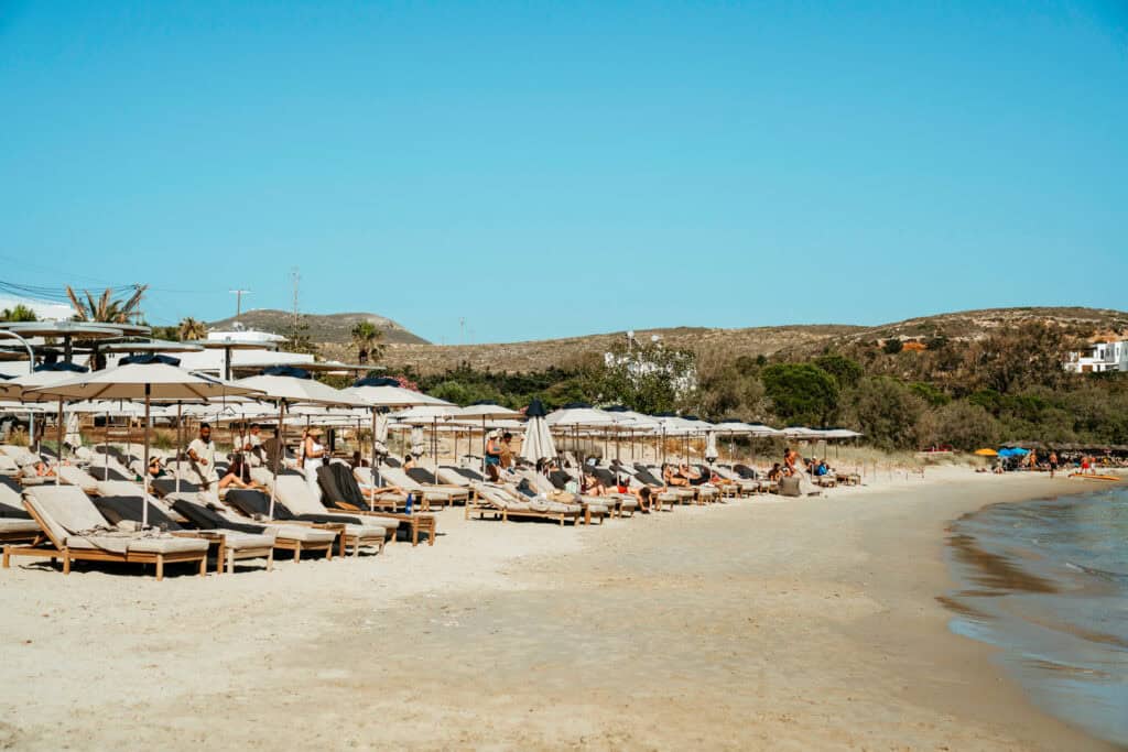 Long stretch of shoreline lined with sunbeds and umbrellas at Marcello Beach Paros, where visitors relax under the sun with views of the surrounding hills.