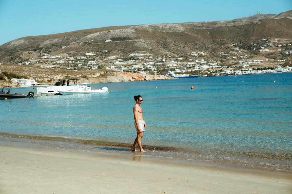 Caleb walking along the shoreline at Marcello Beach Paros with clear turquoise water and boats anchored nearby, offering a view of Parikia across the bay.