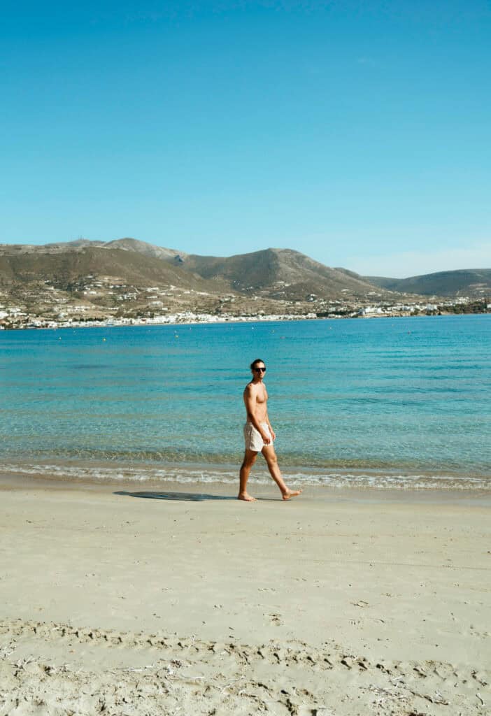 Caleb walking barefoot along the shore at Marcello Beach Paros with clear turquoise water and mountain views in the background.