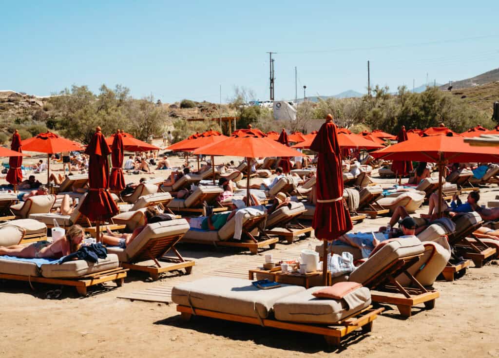 Crowds of sunbathers relaxing on padded loungers under red umbrellas at Monastiri Beach, Paros, with a sandy beachfront and dry hillside landscape in the background.