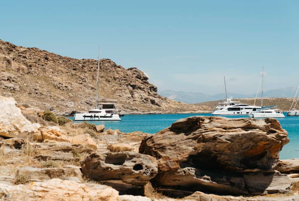 Sailboats and catamarans anchored in the turquoise bay near Monastiri Beach, Paros, with rugged rocky hills and weathered shoreline in the foreground.