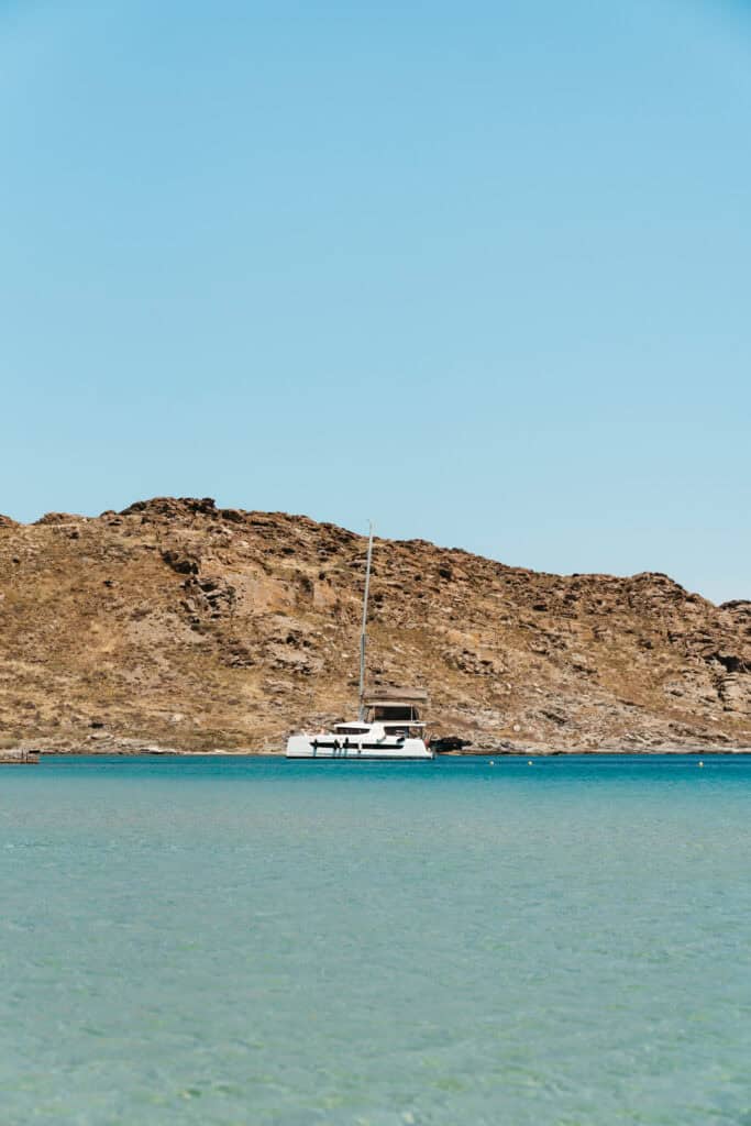 Single white catamaran anchored near the rocky shoreline of Monastiri Beach, Paros, surrounded by calm turquoise waters and set against a backdrop of rugged hills and clear blue skies.