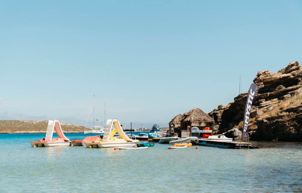Colorful pedal boats with slides floating in shallow water near thatched-roof watersports huts at Monastiri Beach, Paros, with sailboats and rocky cliffs in the background.