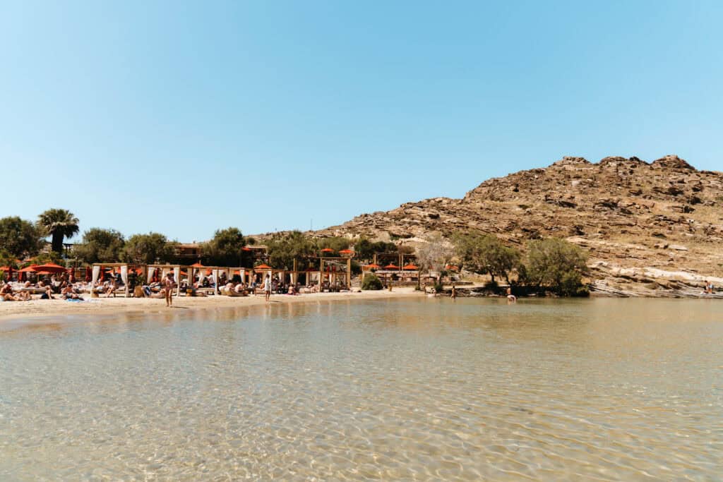 Beachgoers lounging under red umbrellas and shaded cabanas at Monastiri Beach, Paros, with shallow clear water 