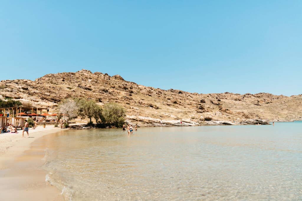 Peaceful stretch of shoreline at Monastiri Beach, Paros, with visitors wading in the shallow water and rocky hills rising behind a beachside lounge area with red umbrellas.
