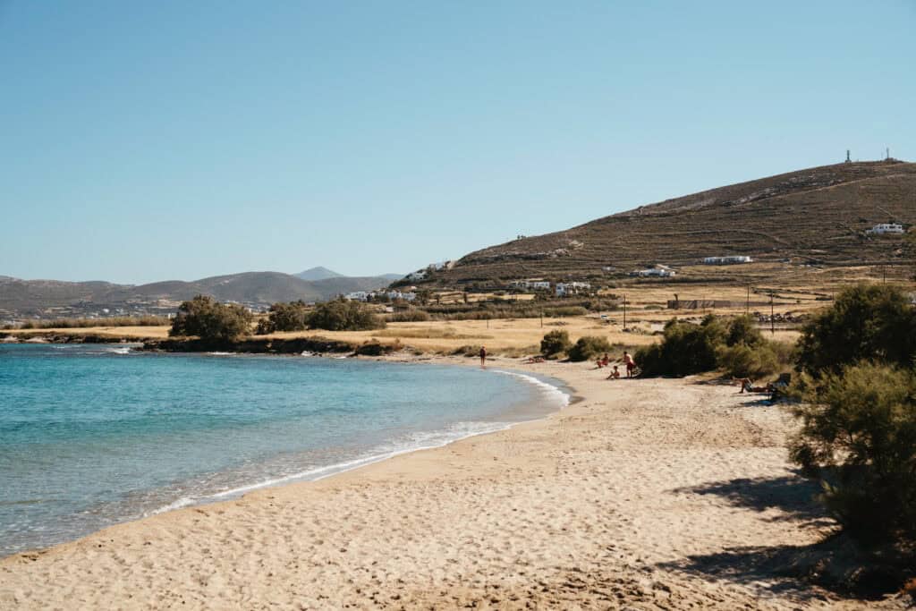 Quiet sandy stretch near Monastiri Beach, Paros, with a few people relaxing along the shoreline, turquoise water lapping the coast, and rolling hills and farmland in the background.