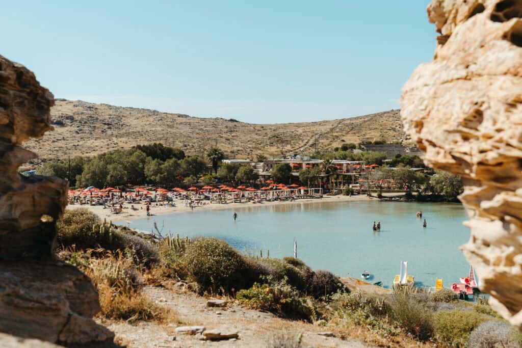 Scenic view of Monastiri Beach, Paros, framed by rocky cliffs, showing turquoise water, rows of red umbrellas and beach cabanas, and pedal boats along the shoreline.