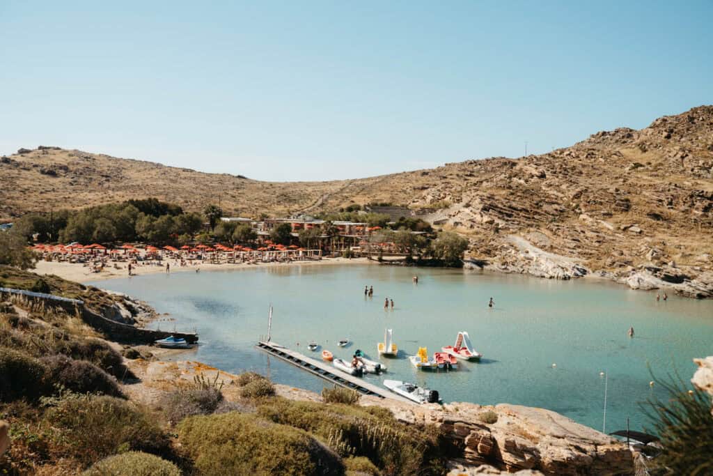 Aerial view of Monastiri Beach in Paros with red umbrellas lining the sandy shore, clear shallow waters filled with paddle boats and swimmers, and rocky hills surrounding the bay.