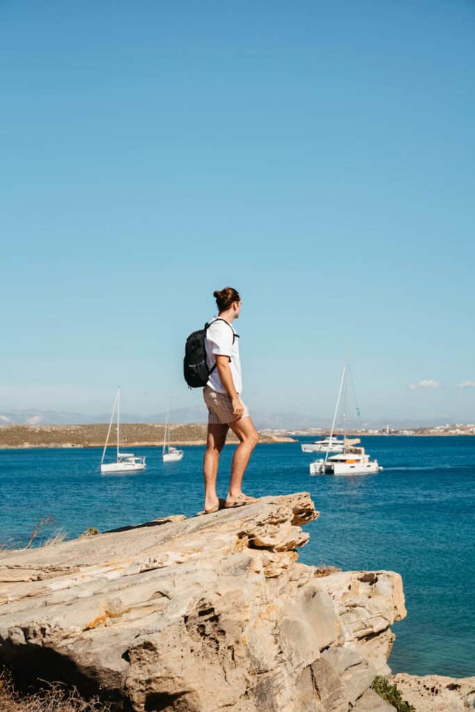 Caleb standing on a rocky cliff overlooking the Aegean Sea near Monastiri Beach, Paros, with sailboats anchored in the turquoise water and mountains in the distance.