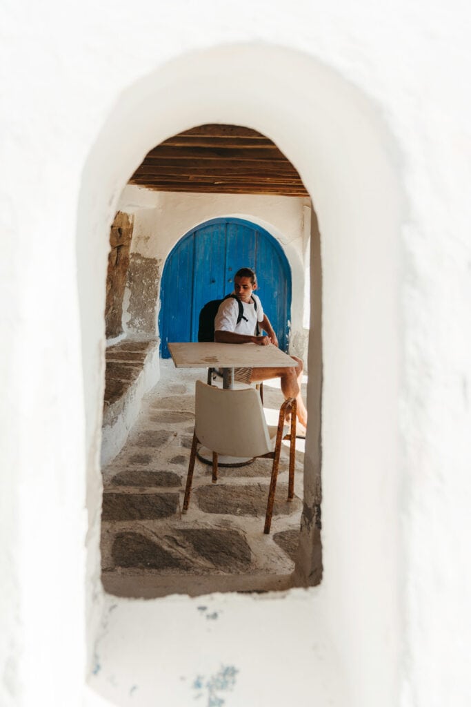 Caleb sitting at a small table in a shaded stone hallway, framed by a white arched entrance and a rustic blue wooden door near Monastiri Beach, Paros.
