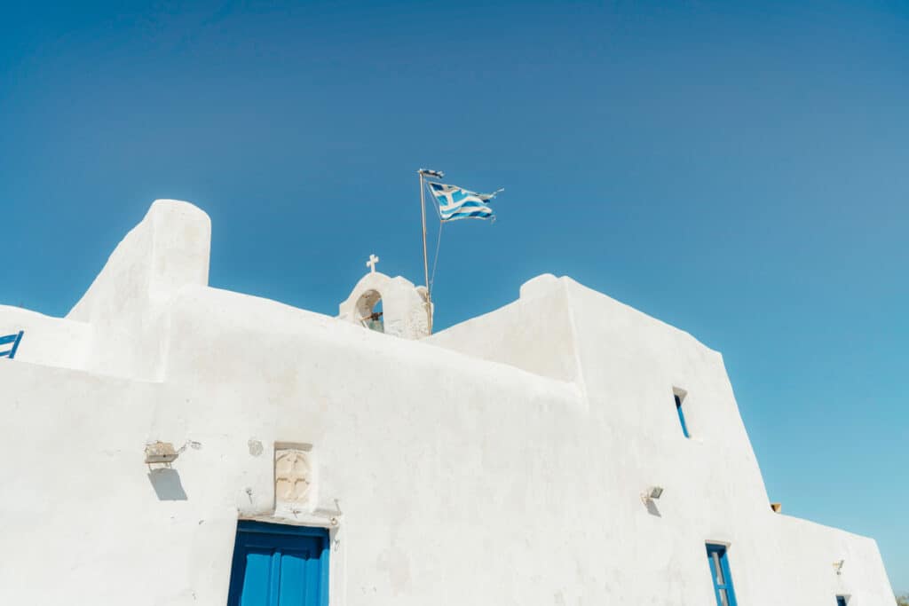 Monastery of St. John's of Deti, a minimalist whitewashed Greek chapel with blue doors and a weathered Greek flag fluttering above, set against a cloudless blue sky near Monastiri Beach, Paros.
