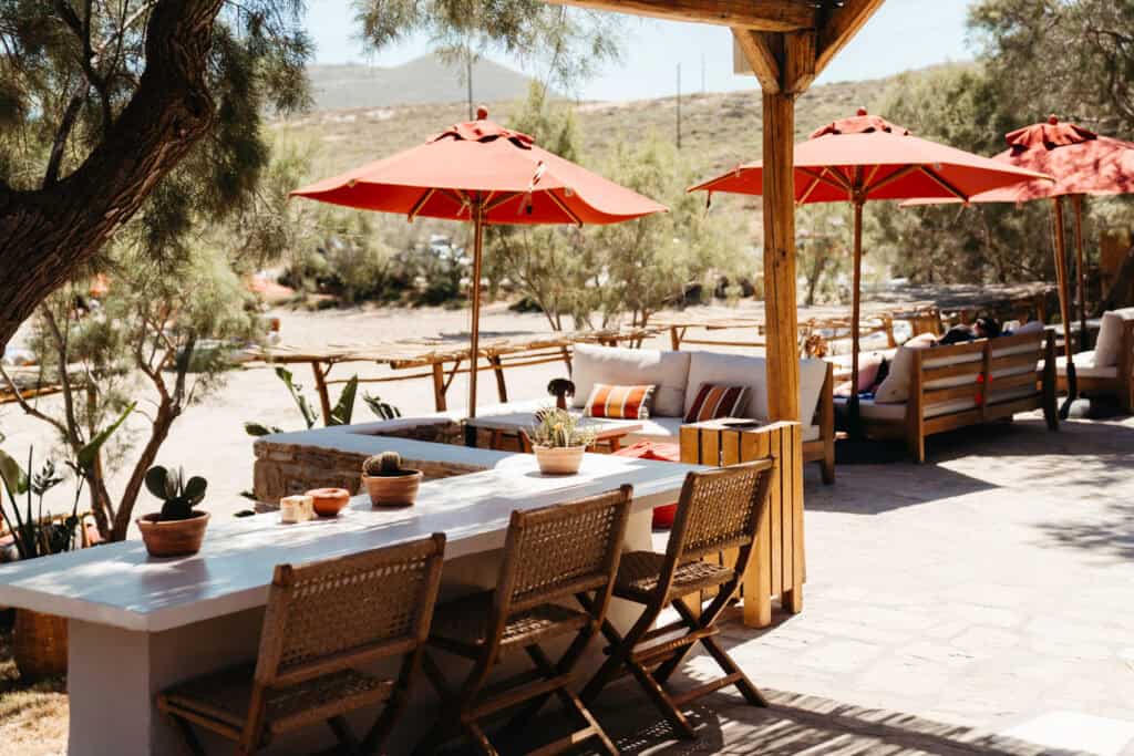 Outdoor lounge area at Monastiri Beach in Paros, featuring wooden chairs, white tables with potted plants, and shaded seating under red umbrellas overlooking a sandy beachfront.