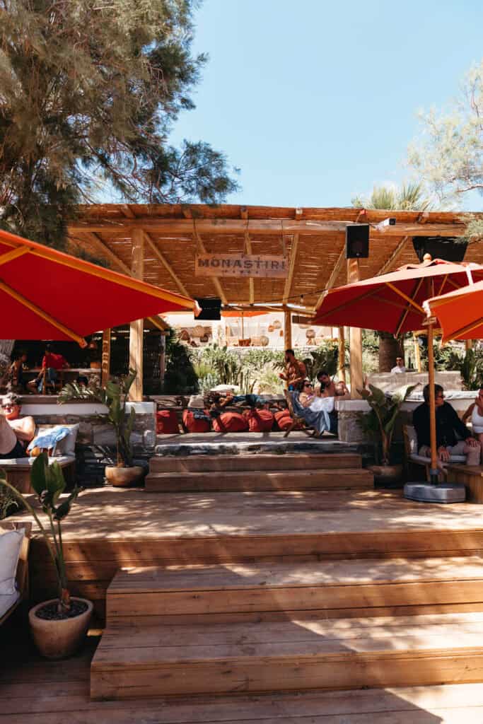 Entrance to the Monastiri Beach Club in Paros, with wooden steps leading up to a shaded lounge area featuring red cushions, potted plants, and people relaxing under red umbrellas.