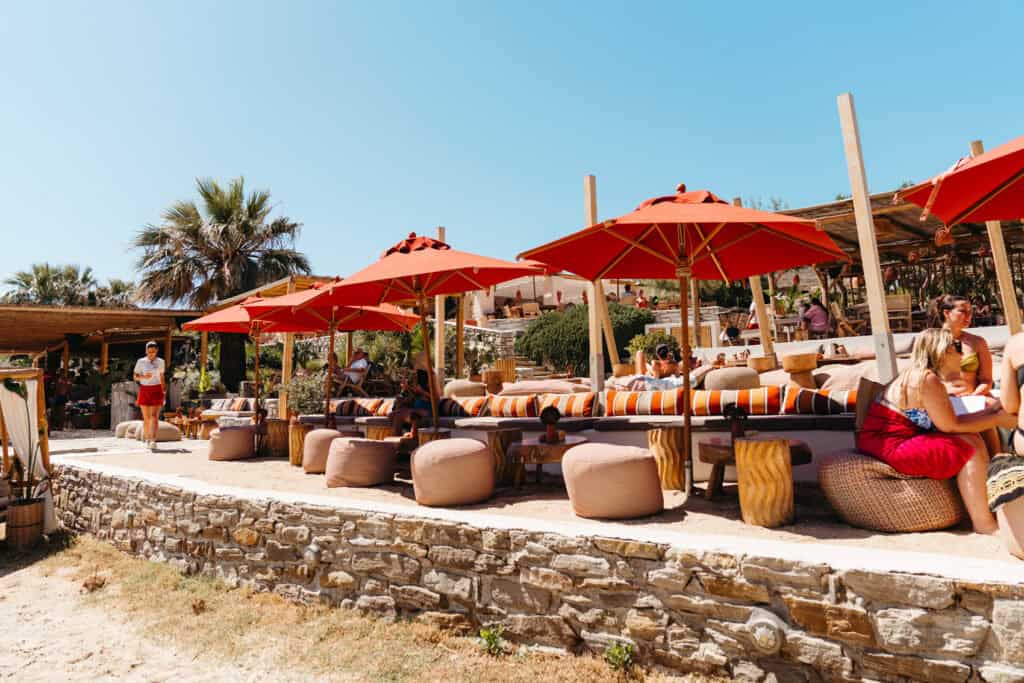 Outdoor lounge area at Monastiri Beach, Paros, with striped cushions, poufs, and red umbrellas set against a stone wall and palm trees, creating a stylish and relaxed beach club atmosphere.