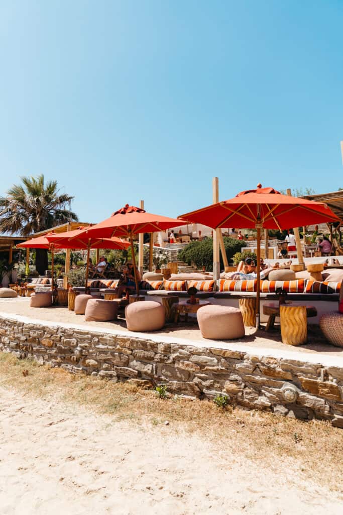 Outdoor lounge area at Monastiri Beach, Paros, with striped cushions, poufs, and red umbrellas set against a stone wall and palm trees