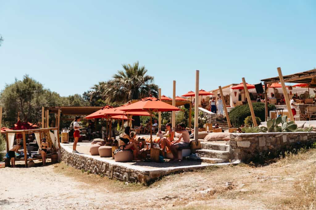 Guests relaxing under red umbrellas at Monastiri Beach Club in Paros, surrounded by palm trees, rustic stone terraces, and cushioned lounge seating near the sandy shore.